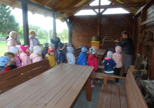 Grupa dzieci zebrana pod drewnianą wiatą wraz z panią dyrektor, panią Agnieszką i p. Basią słucha pani opowiadającej o narzędziach pracy pszczelarza.